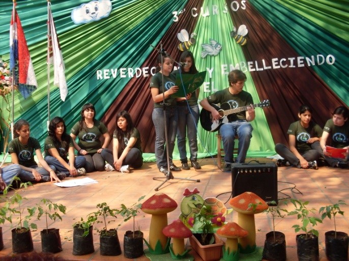 A group of people sitting on a stage in front of plants, embelleciendo the environment through reverdeciendo efforts.