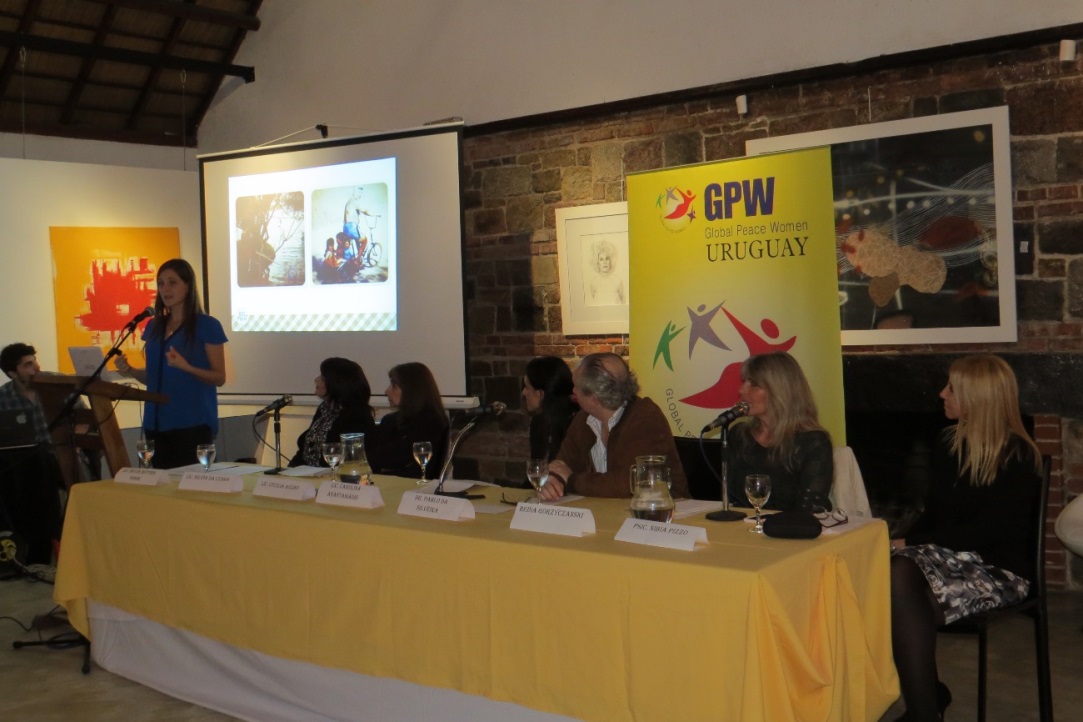 A speaker presenting at a panel event with a projected image, audience, and panel members seated at a table with name placards on Women's GPW Testimonies.