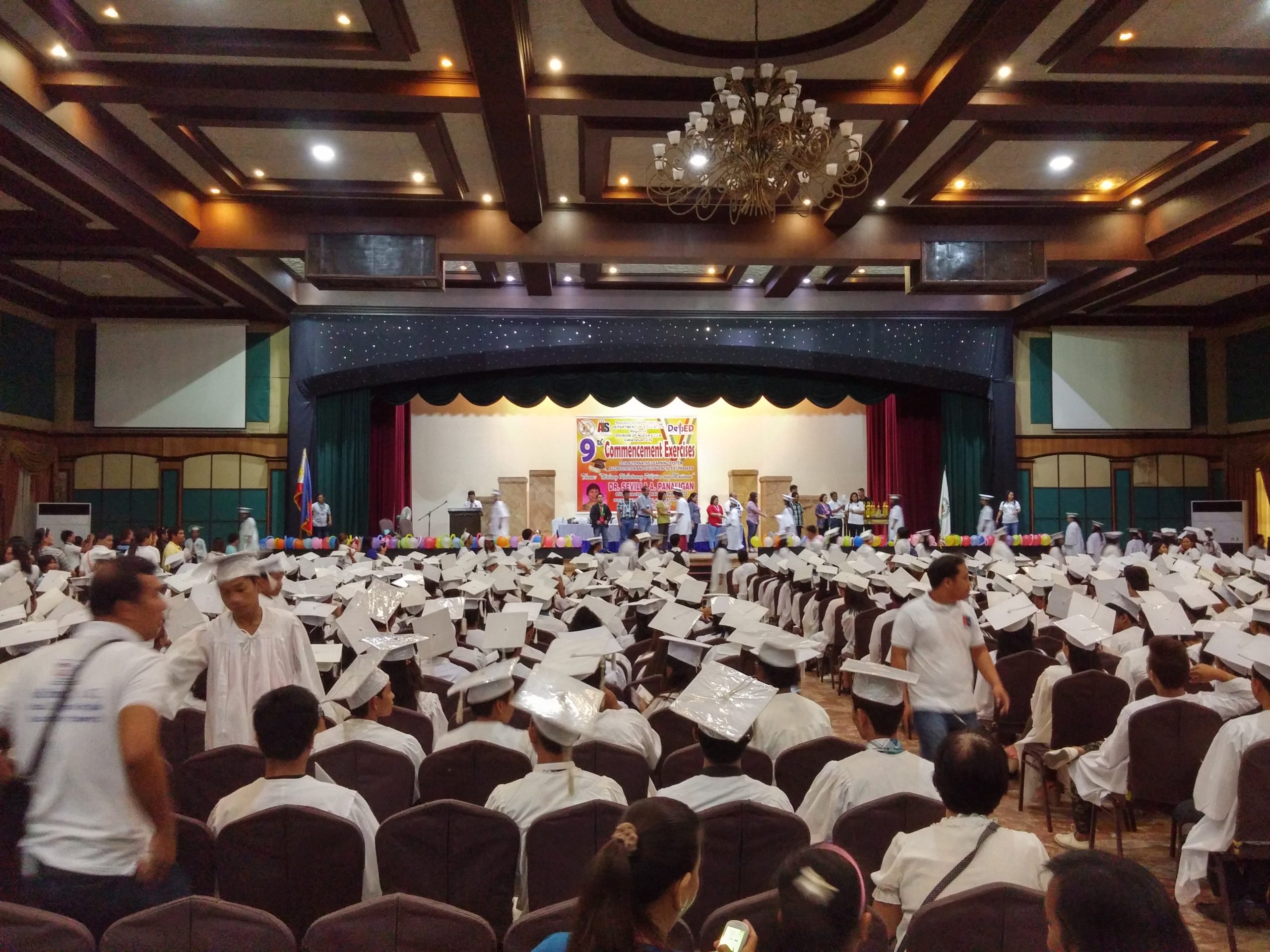 Graduation ceremony in progress with students in white caps and gowns seated facing a decorated stage.