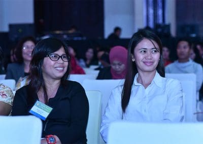 Two women smiling and sitting among an audience at an indoor event.