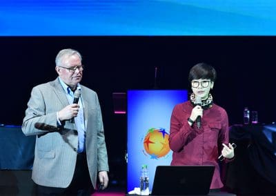 Two speakers presenting at a conference with a digital screen in the background.
