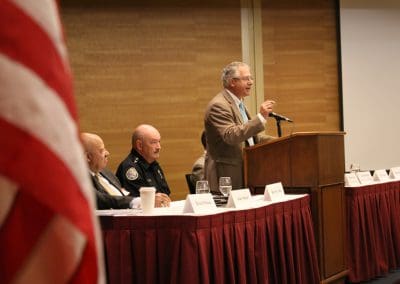 A man speaking at a podium during a panel discussion with two seated individuals listening.