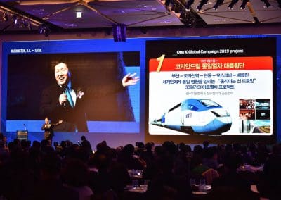 A speaker presents at an event, with a large screen displaying their content and image behind them.