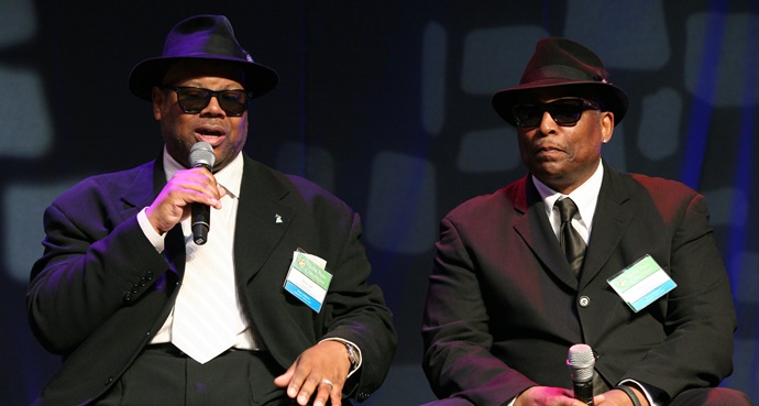 Two men in suits and hats speaking at an event, one holding a microphone.