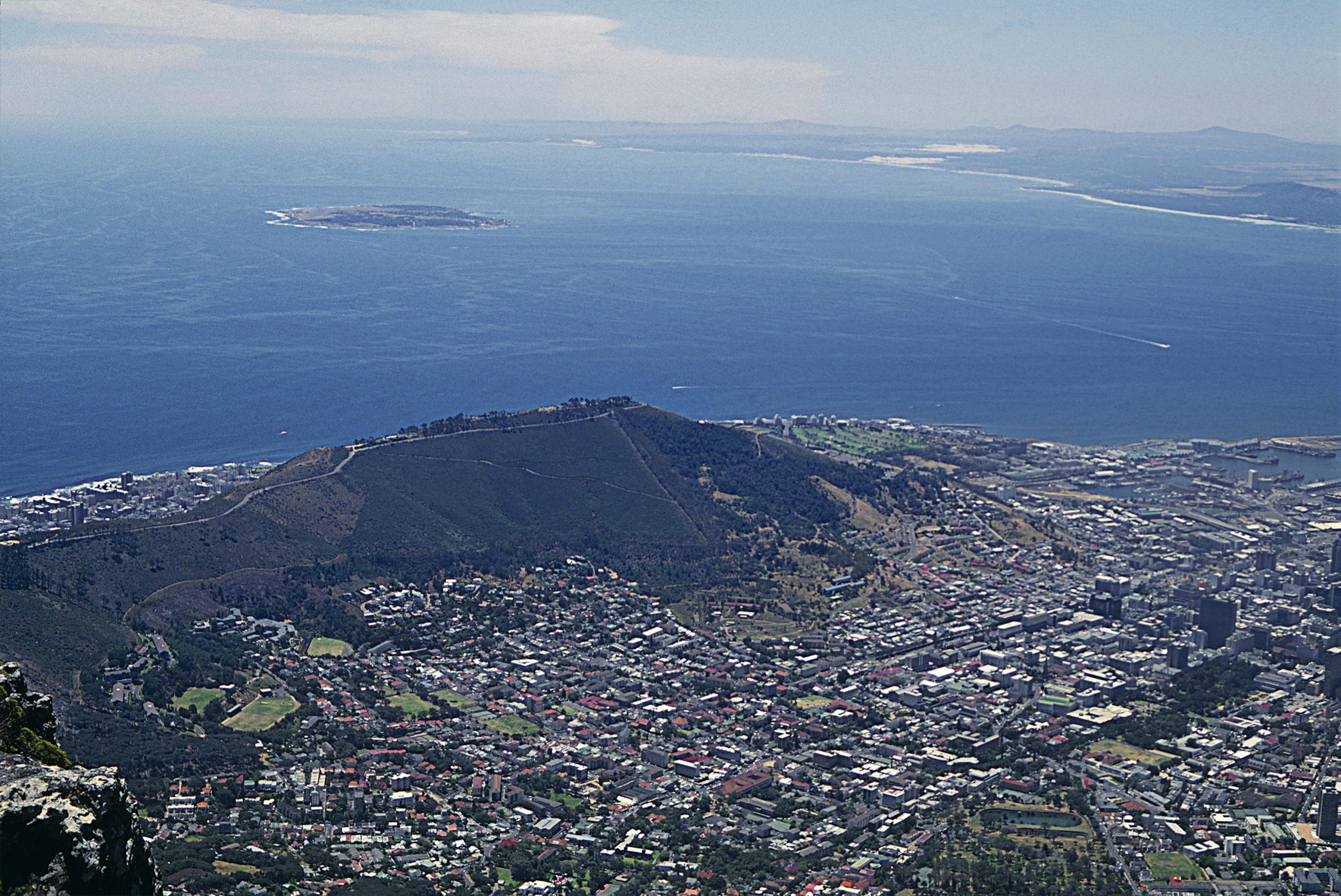 Aerial view of a coastal city with dense buildings leading up to a green hillside and an expansive view of the ocean in the background.