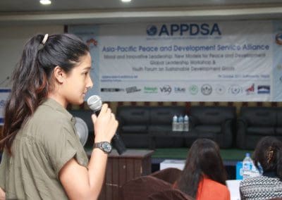 A woman speaking into a microphone at a forum with an audience in the background.