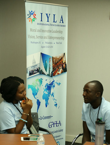 Two individuals engaged in conversation at an event with a banner for the international young leaders assembly (iyla) in the background.