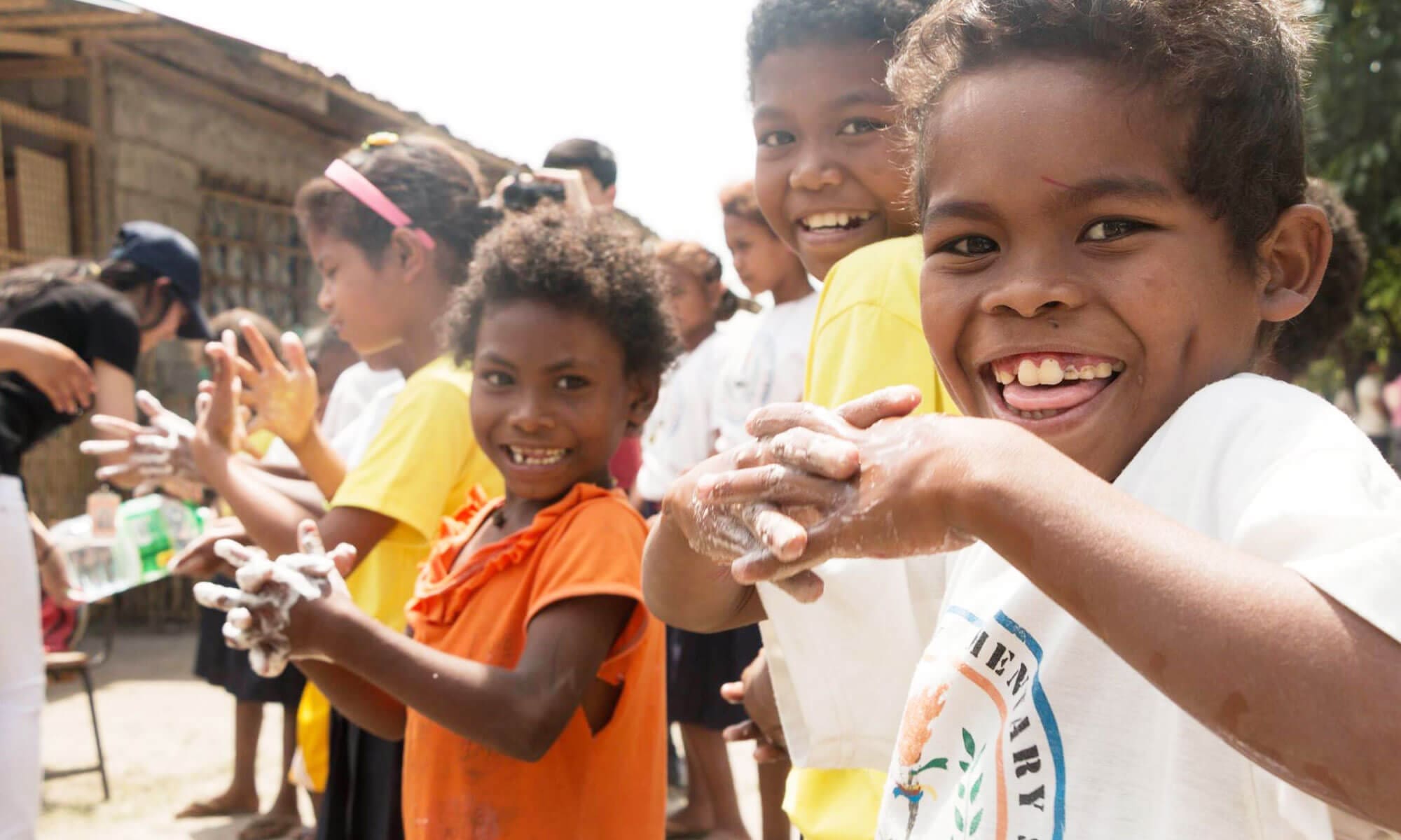 A group of people gather around and inside a shallow pit filled with water, using metal buckets to collect and distribute the water.
