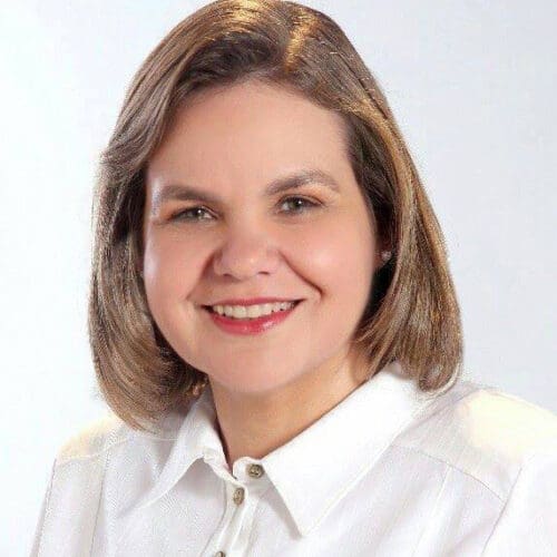 A person with shoulder-length hair, reminiscent of a Paraguayan Senator, smiles while wearing a white collared shirt against a light background.