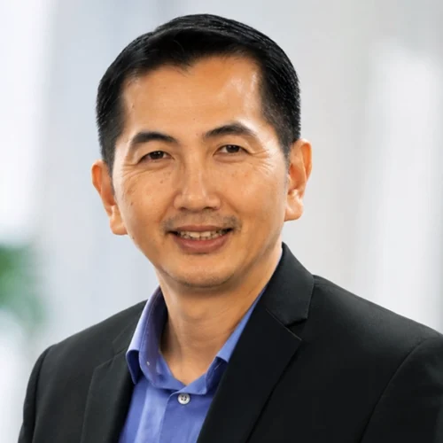 Su Thye Teh, dressed in a dark suit and blue shirt, smiles at the camera against a blurred indoor background.