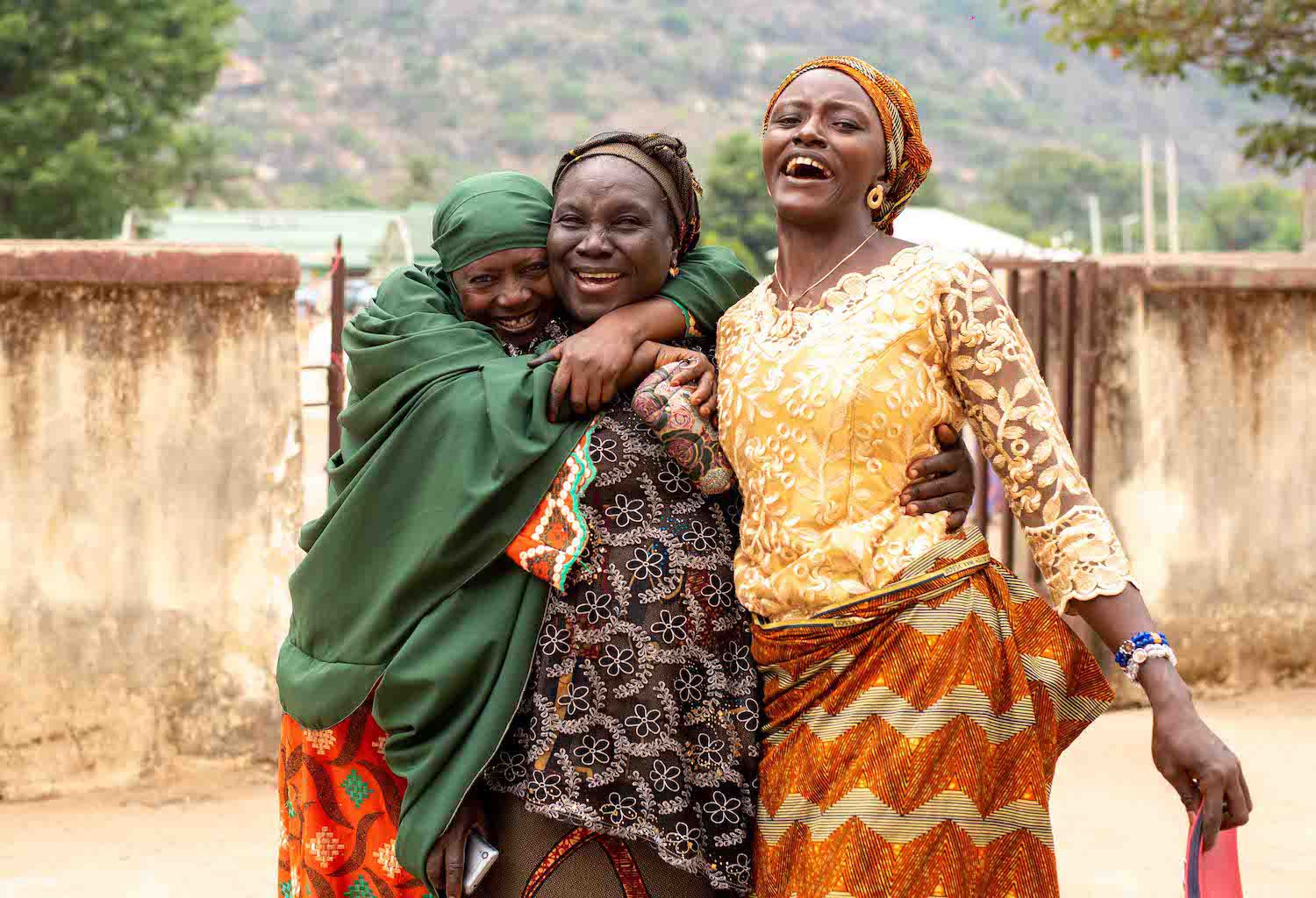 Three African women joyfully embracing on a street, with one woman laughing and the others smiling broadly after attending a financial literacy workshop.