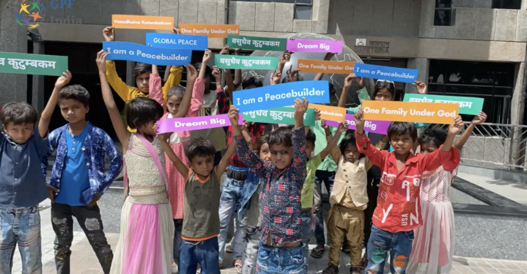 Children from the Vasudhaiva Kutumbakam Learning Center hold sign reading 'I am a Peacebuilder', 'Global Peace', 'Dream Big', and 'One Family Under God'.