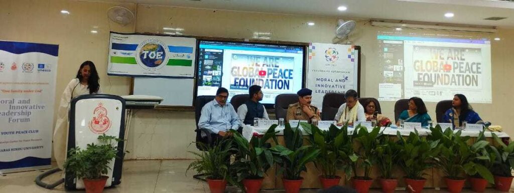 A group of people sitting at a table in front of a screen during the 'Vasudhaiva Kutumbakam' Moral and Innovative Leadership Forum on Peace and Governance held in India.