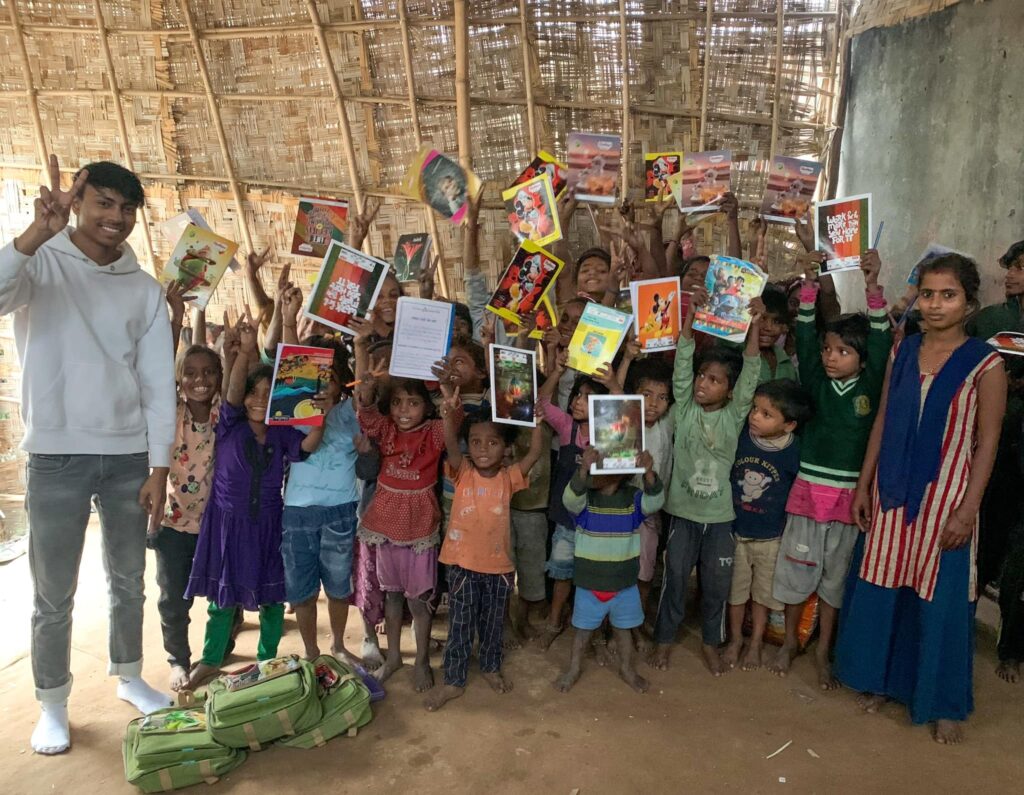 A group of children holding up books in front of a hut during the S.M.I.L.E Annual Day organized by GPF India.