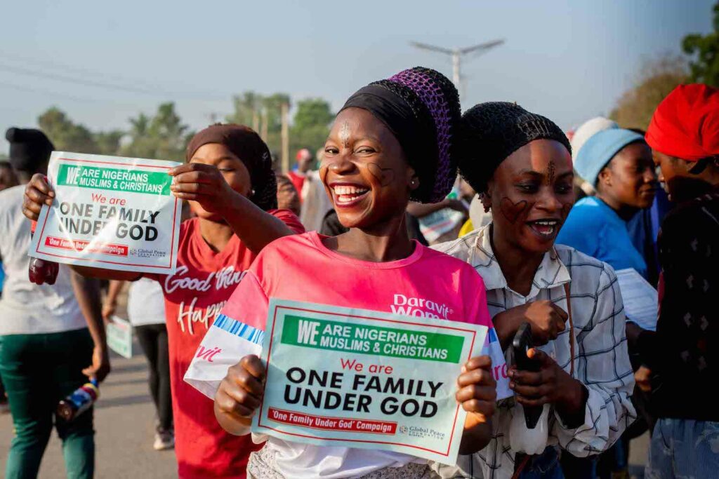 A group of women holding signs that say one family under god, promoting community peace in Nigeria during the Afan Festival.