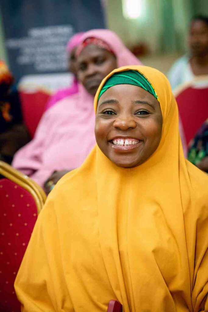 A woman wearing a yellow hijab smiles in front of a group of people, representing the Economic Empowerment and Women's Cooperative Societies in Nigeria supported by GPF Nigeria.
