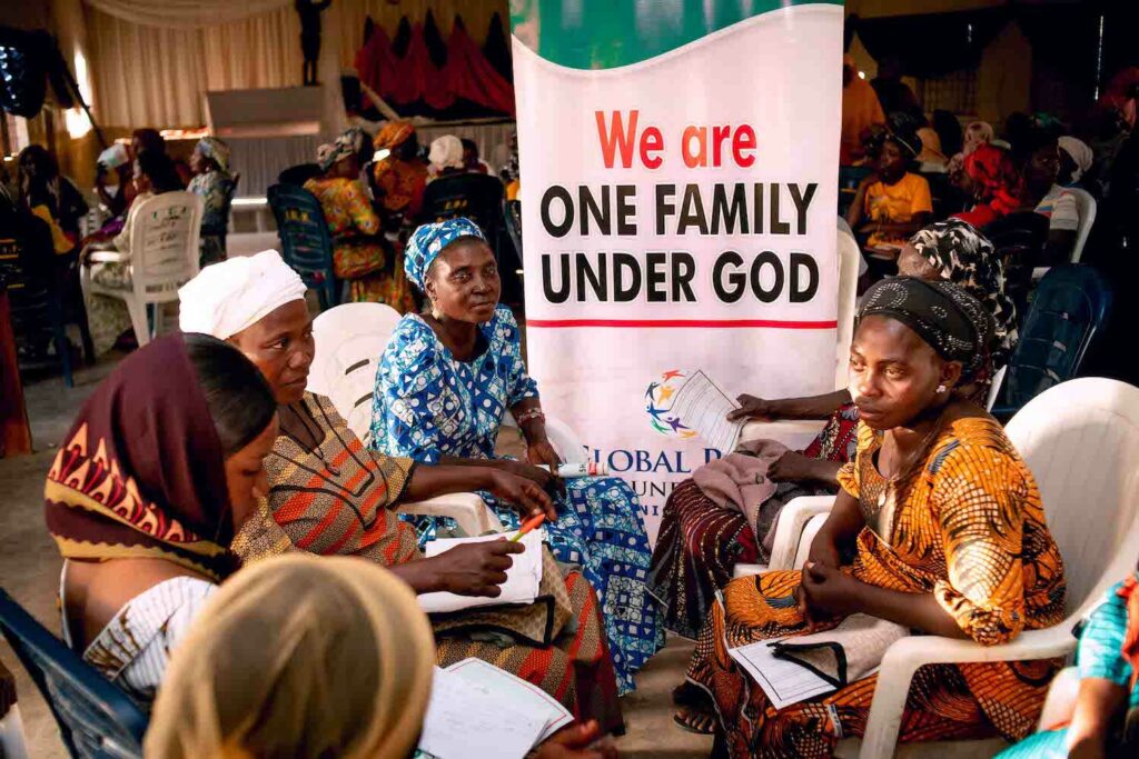 A group of women from Women's Cooperative Societies sitting around a sign that says we are one family under God, promoting Economic Empowerment through GPF Nigeria.