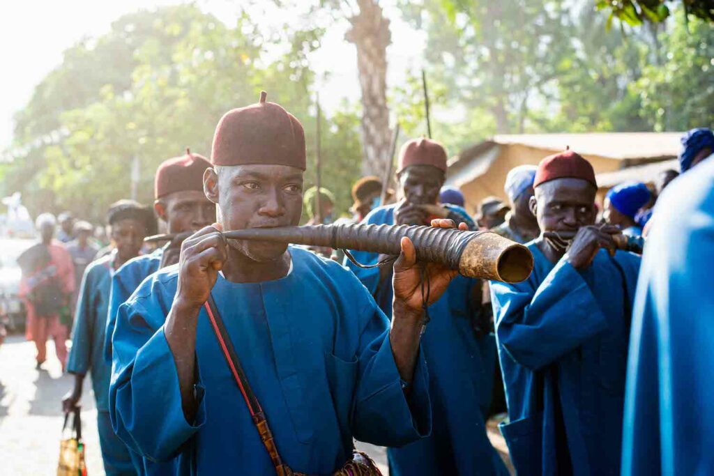 A group of men in blue robes performing at the Afan Festival in Nigeria, playing a flute to foster sustaining community peace.