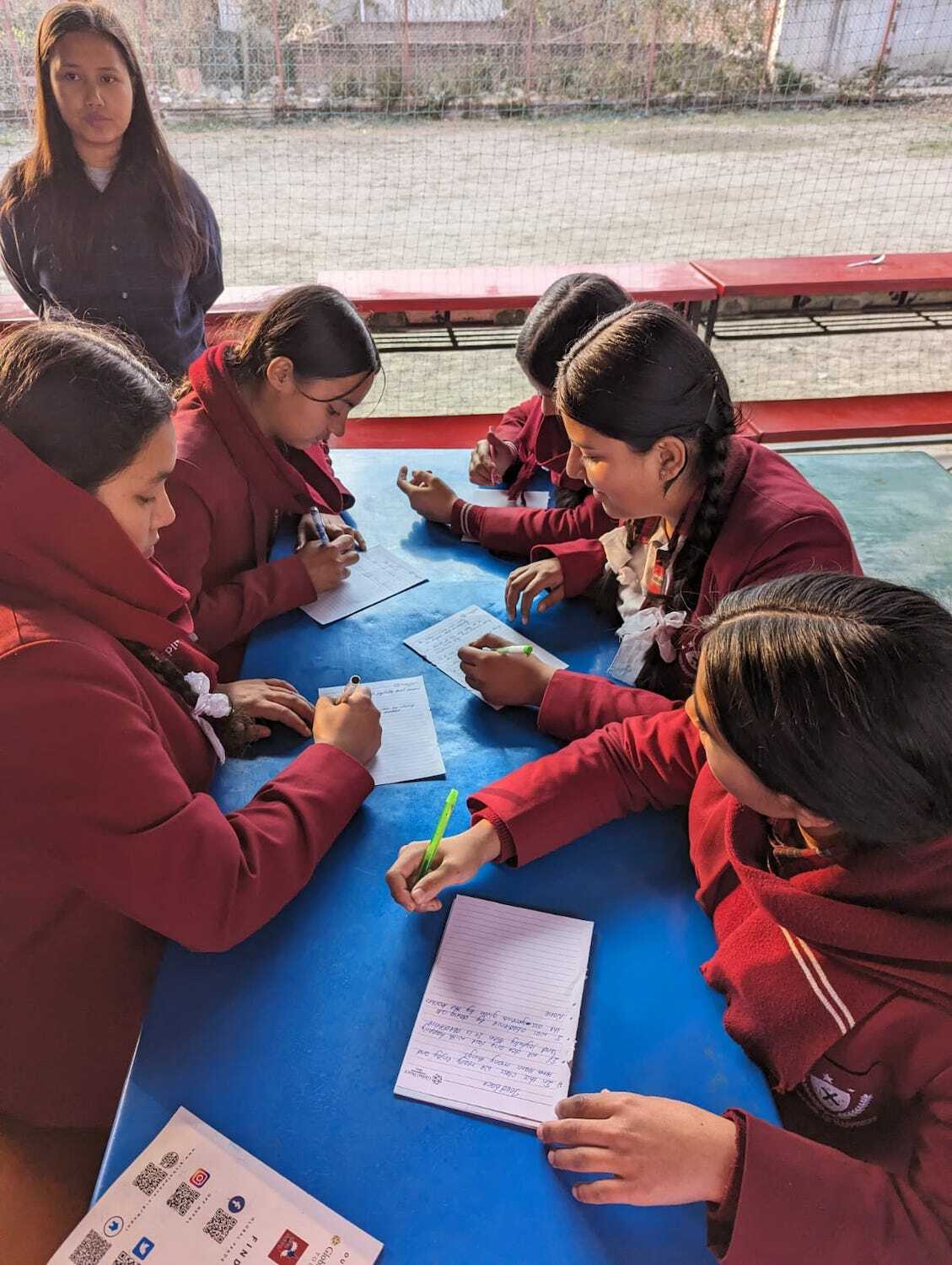 A group of students in red uniforms seated around a table, engaged in writing and discussion on values-based education, with one person standing in the background observing.