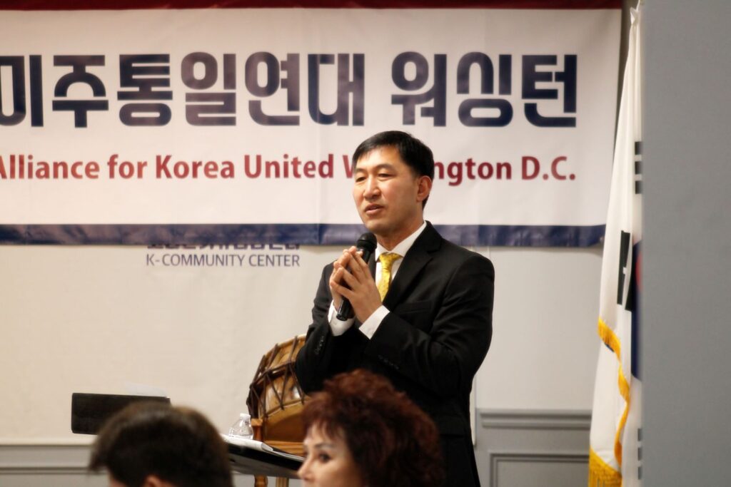 A man speaking at a forum with a banner for the Alliance for Korea United Washington D.C. in the background.