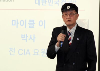 A man in a beret and suit delivering a presentation at the Alliance for Korea United forum with projected Korean text in the background.