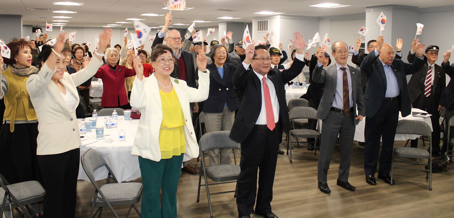 Group of people standing and raising their hands with small flags at the Alliance for Korea United forum in a meeting room.