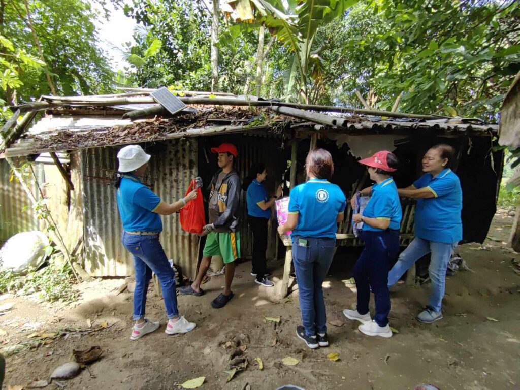 A group of people from the Inner Wheel Club of Central Ortigas, wearing blue shirts with a logo, are engaged in a community service activity, providing assistance or supplies to a resident at a humble