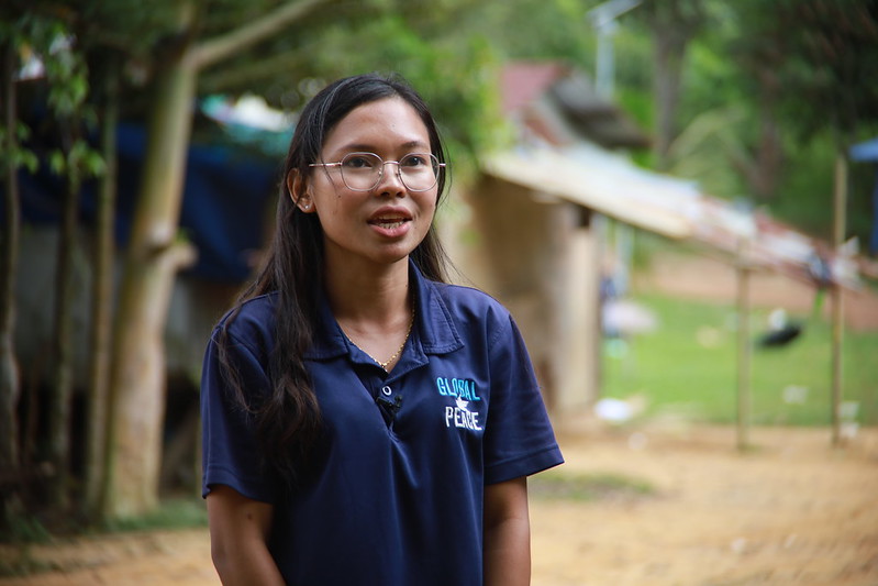 A woman in a blue shirt standing outdoors with trees in the background of a remote village.