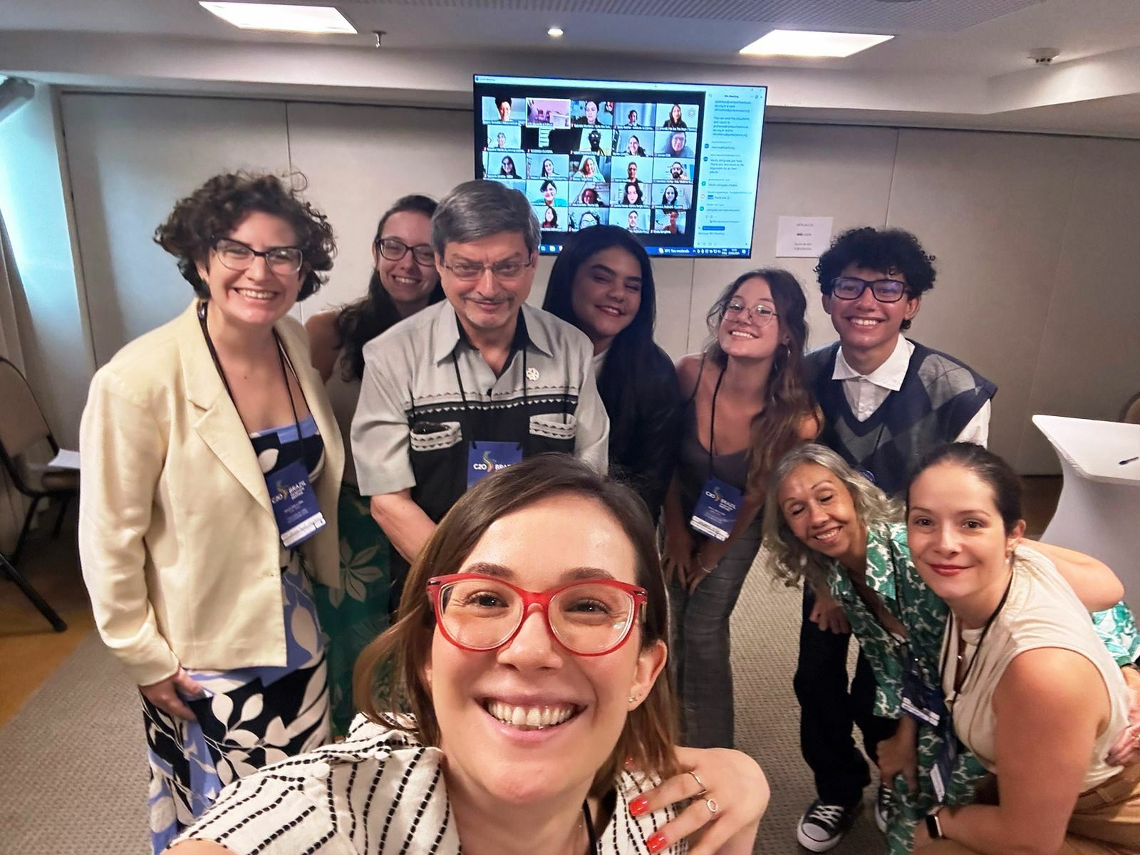 Group of nine cheerful people taking a selfie at the "C20-2024 Brazil" education and culture session, with a screen displaying additional participants in the background.