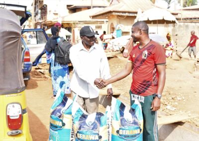 Two men shaking hands in a busy market street in Kagoro, with sacks of grain and a yellow car in the background.