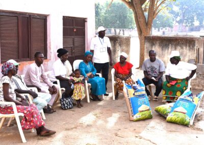 Group of adults and a child sitting outdoors with bags of rice, interacting with a standing man wearing a badge, in Kagoro.