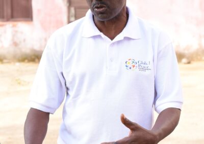 A man in a white polo and black cap stands talking about humanitarian aid, with one hand slightly raised, outdoors during the day.