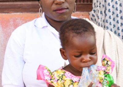 A woman in a white shirt and black headscarf sitting with a toddler in a floral dress, who is drinking from a bottle in Kagoro.