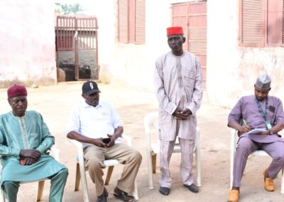 Four elderly men in traditional and casual attire sitting and standing in a Kagoro courtyard, engaging in a discussion.