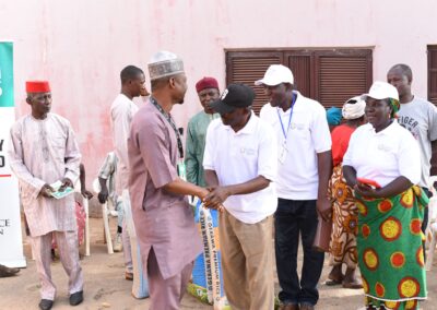 Two men shaking hands at an outdoor community event in Kagoro, with other attendees watching, standing near banners promoting health and peace.