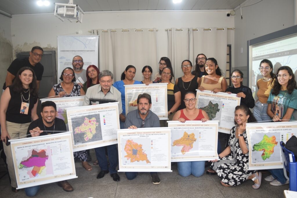 Group of Brazilians smiling and holding certificates with maps, in a classroom with a projector screen in the background.