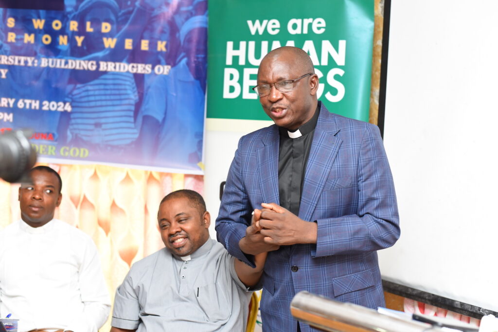 A clergyman addressing an audience during Interfaith Harmony Week in Nigeria, 2024, with banners promoting human unity and diversity in the background.