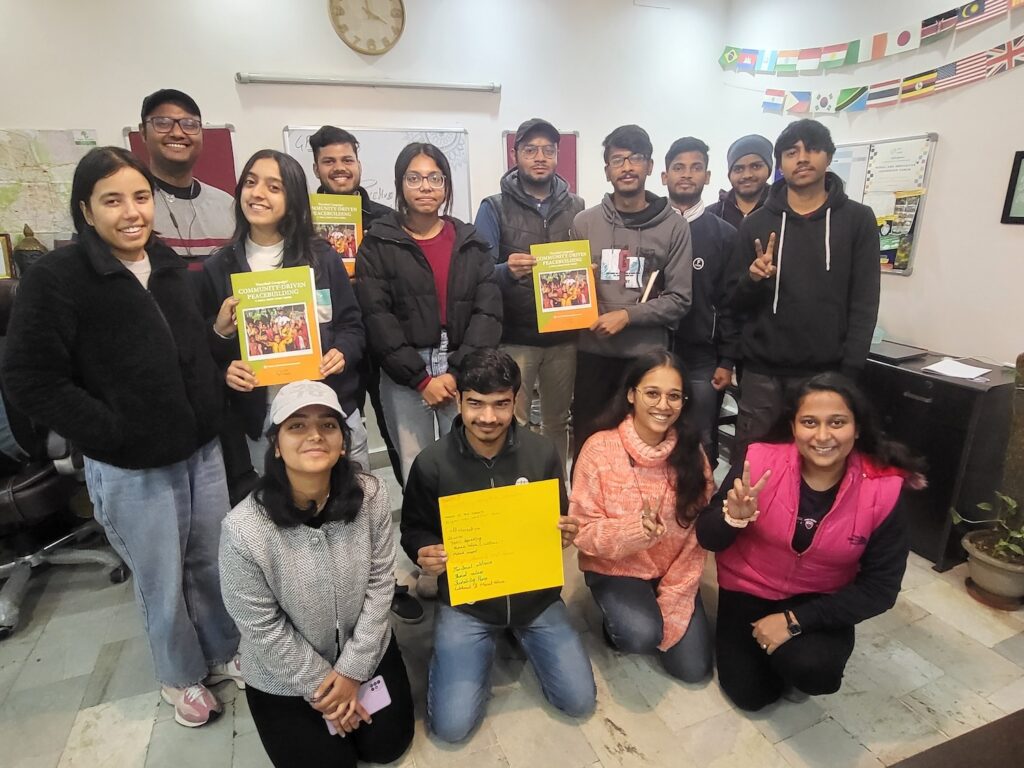 A group of smiling young adults posing for a photo in an indoor setting, some holding books and papers for the PeaceHub Campaign.