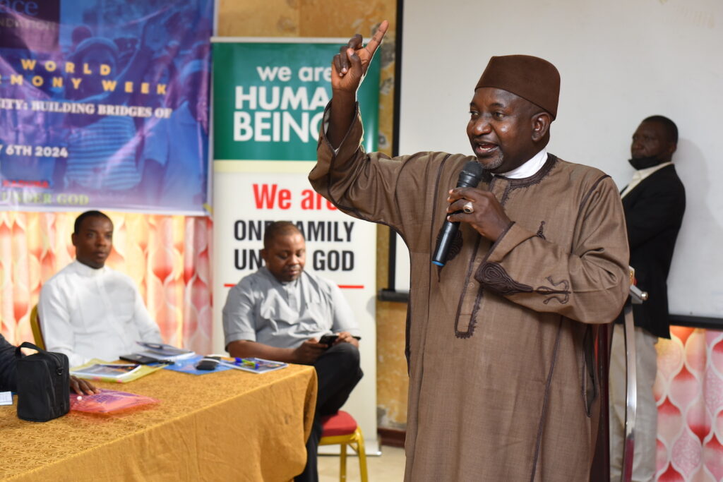 A man in traditional Nigerian attire speaking at a microphone with a banner in the background announcing Interfaith Harmony Week 2024.