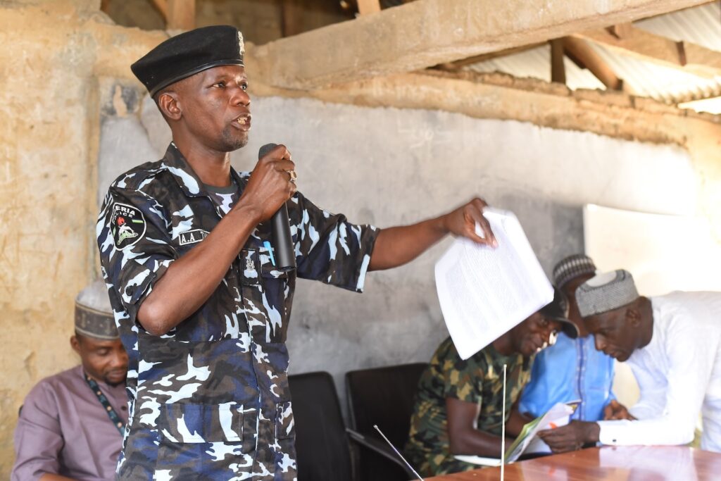 A uniformed officer giving a speech on Peaceful Coexistence and holding papers in a room with seated men listening attentively.