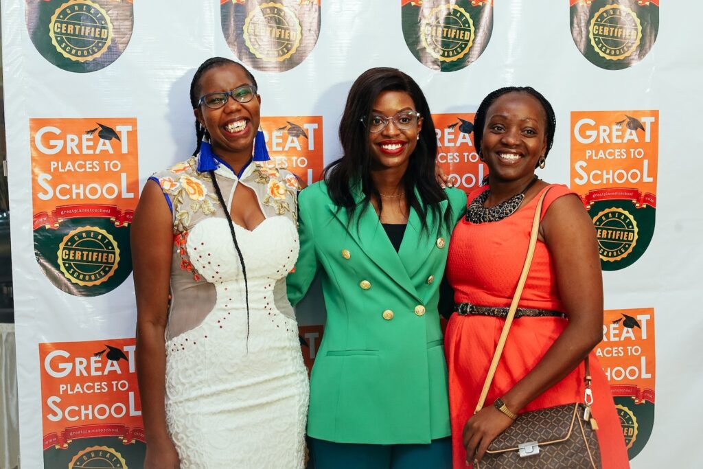 Three women smiling at the 'Outstanding Schools Award' event, standing before a branded backdrop, dressed in smart casual outfits.