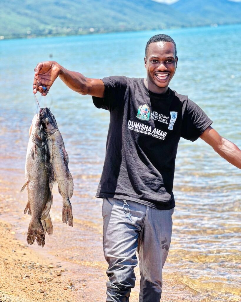 Man holding up two fish by the lakeside with a smile, embodying the spirit of Dumisha Amani among youth peacebuilders.