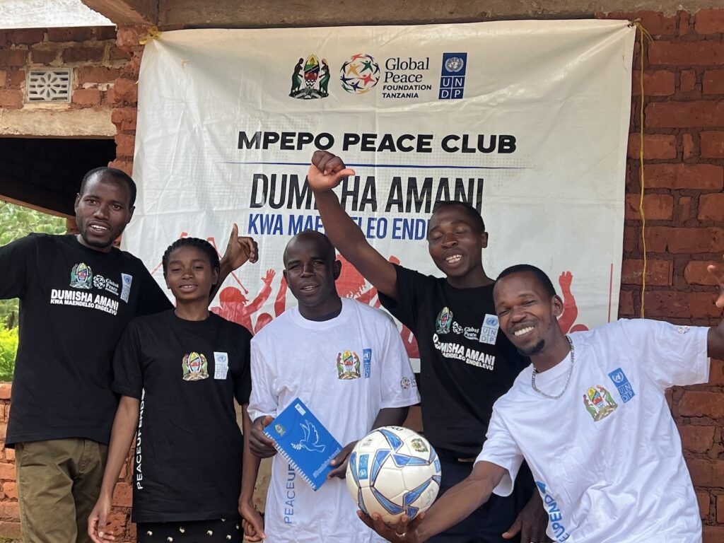 Group of six individuals posing with a soccer ball in front of a banner for Dumisha Amani peace club.