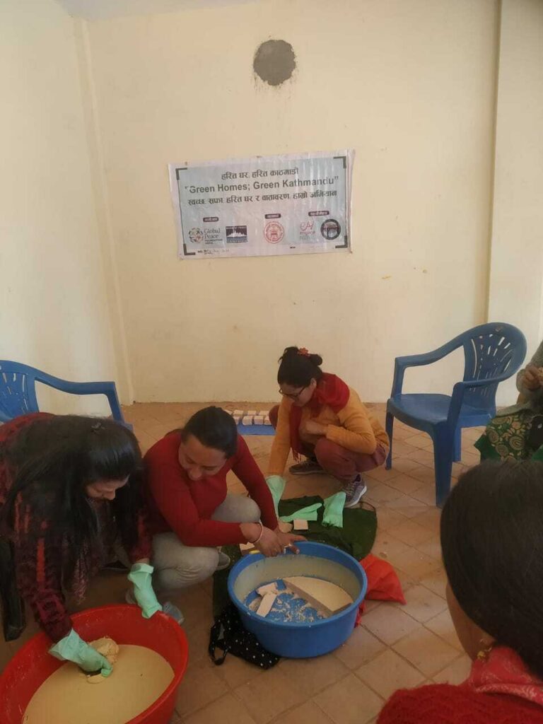 Three women in a room in Nepal, wearing gloves and working with environmentally-friendly products in plastic basins, under a banner about 'green homes, green Kathmandu.'