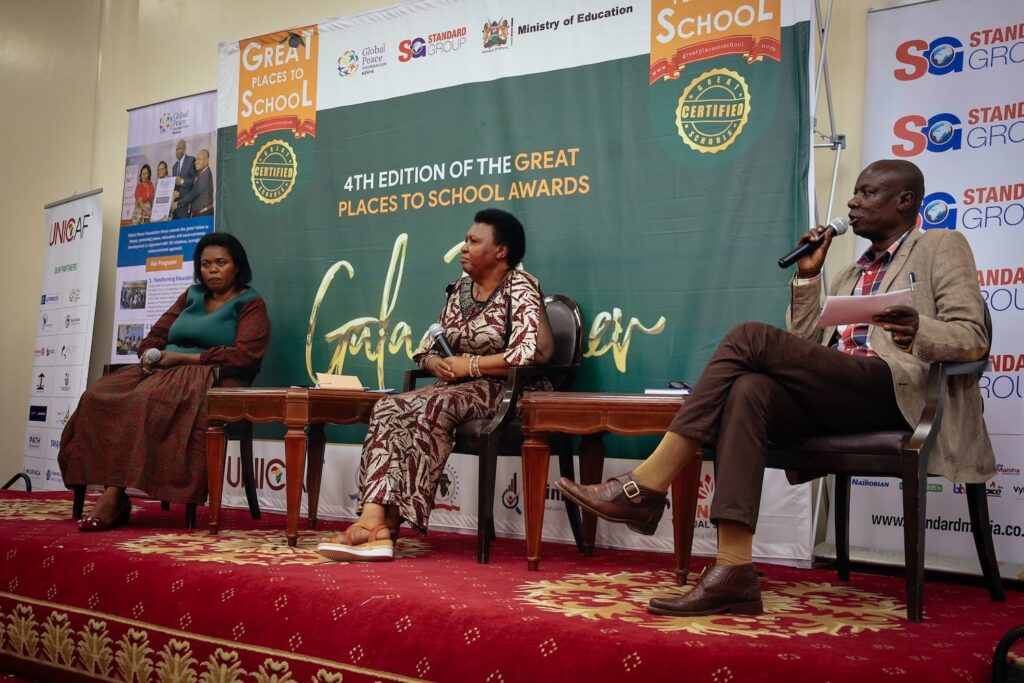 Three panelists sitting on stage during a conference presentation, with banners promoting Holistic Learning in the background.