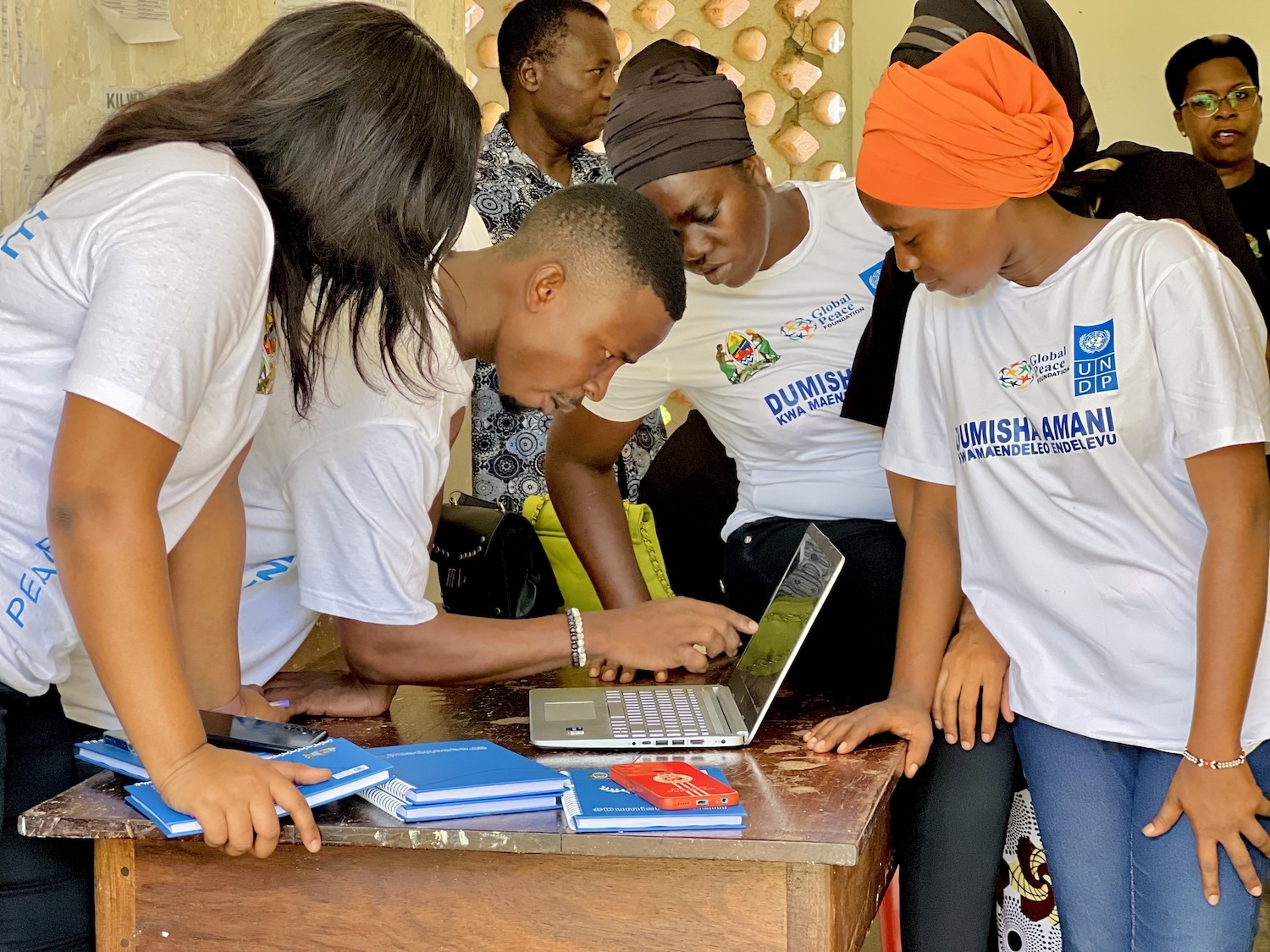 Group of focused individuals in matching t-shirts gathered around a laptop, working as Peacebuilders in Tanzania.