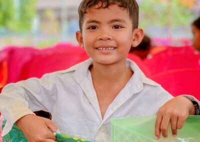A young boy in a white shirt smiles while holding a green folder and a packaged T-shirt under a colorful tent, all part of the Annual Campaign.