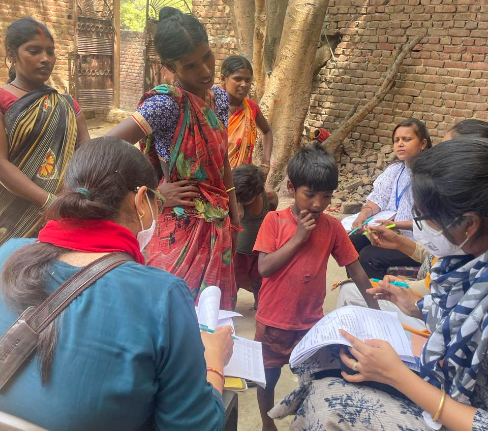 A group of women and children stand while healthcare volunteers, seated with paperwork and pens, engage with them in an outdoor setting near a brick wall and tree at the Vasudhaiva Kutumbakam Learning Center in India.