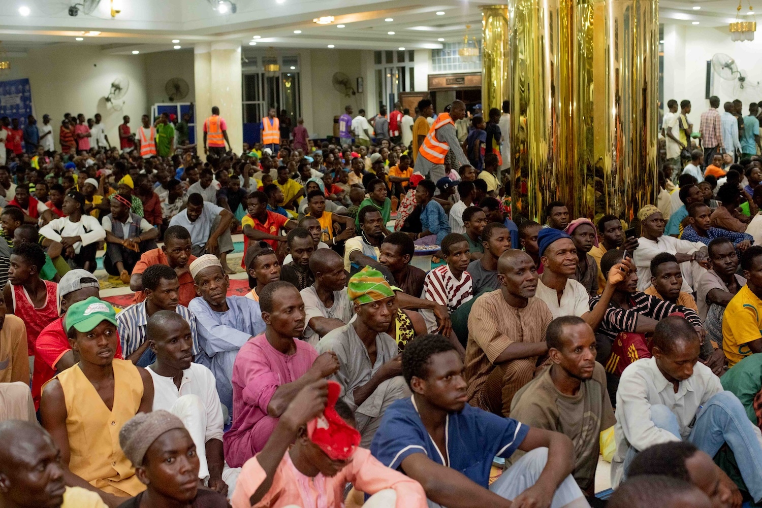 A crowded hall in Nigeria with many seated people, some wearing traditional and reflective vests, attending an event, focusing attentively forward.