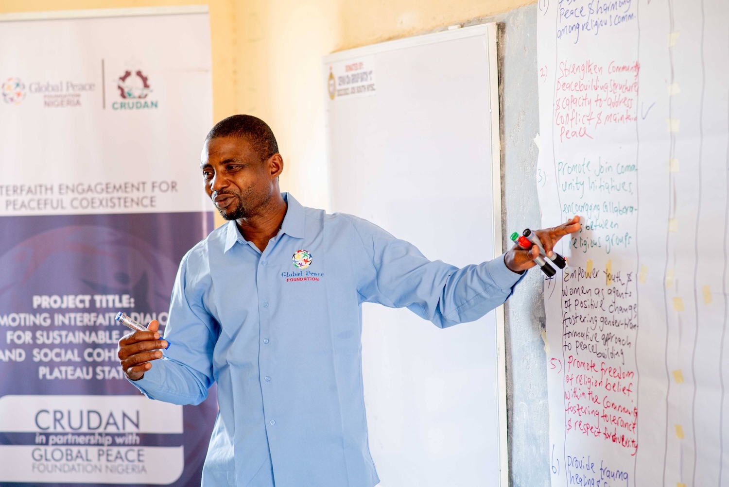 A man presenting at a workshop points to a flip chart with text about interfaith dialogue and sustainable peace.